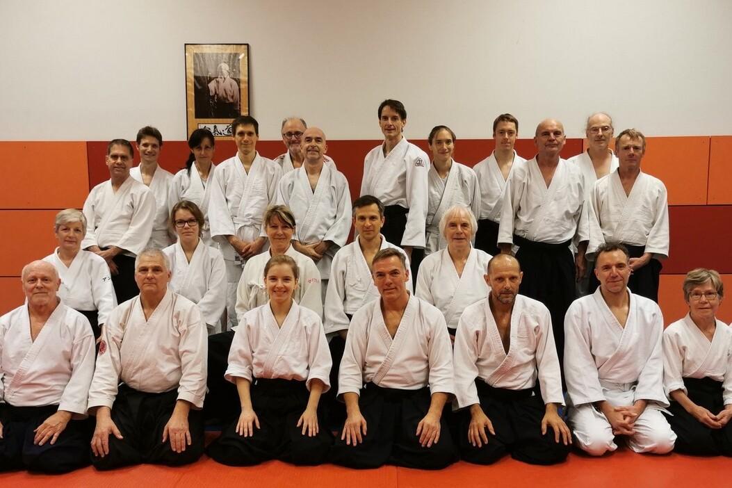 Gruppenfoto von 24 Personen in wei&szlig;en Aikido-Anz&uuml;gen, sitzend und stehend auf einer blauen und roten Matte.