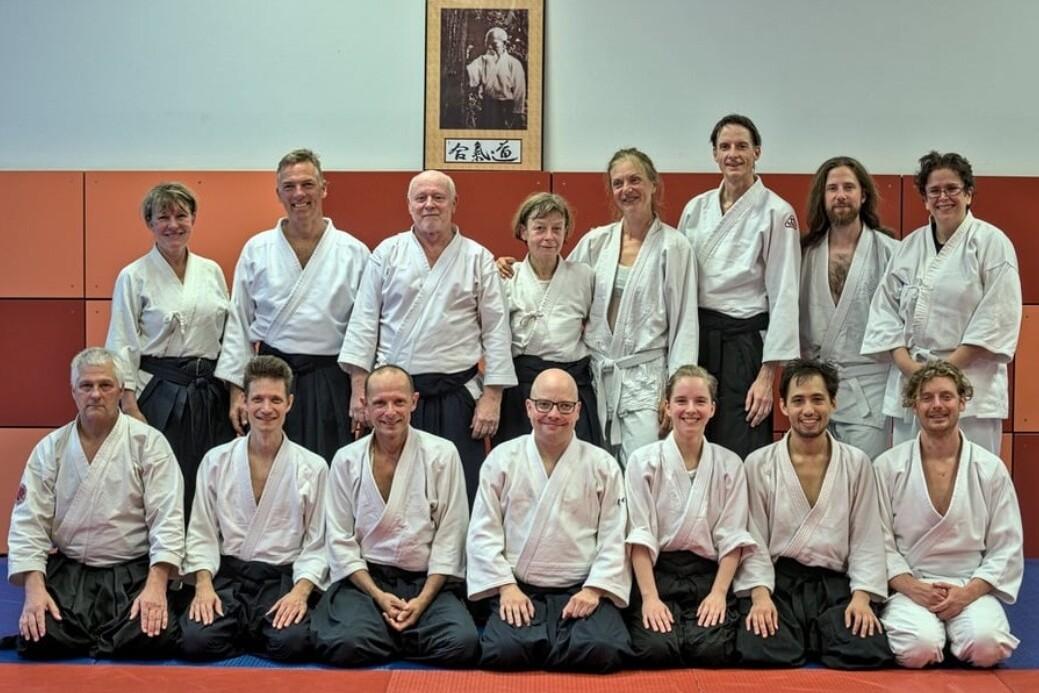 Gruppenzusammenstellung von 15 Menschen in traditionellen Aikido-Uniformen, arrangiert vor einer Wand mit Bild.