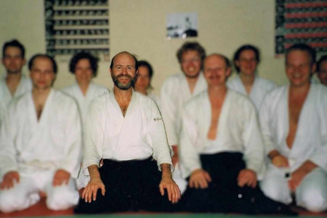 Gruppennahaufnahme von zehn Menschen in wei&szlig;en Judogi, sitzend und stehend in einer Trainingsumgebung.