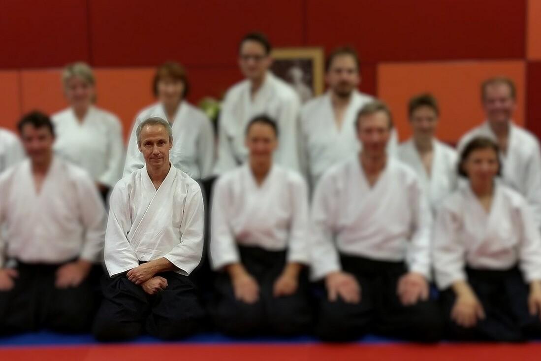 Gruppenfoto von Aikido-Praktizierenden in wei&szlig;en Outfits auf einer blauen Matte, der Fokus liegt auf dem sitzenden Trainer.