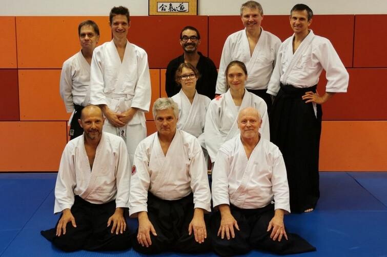 Gruppenshot von zehn Aikido-Praktizierenden in Uniform, auf einer Matte vor einer wandmontierten Fotografie.