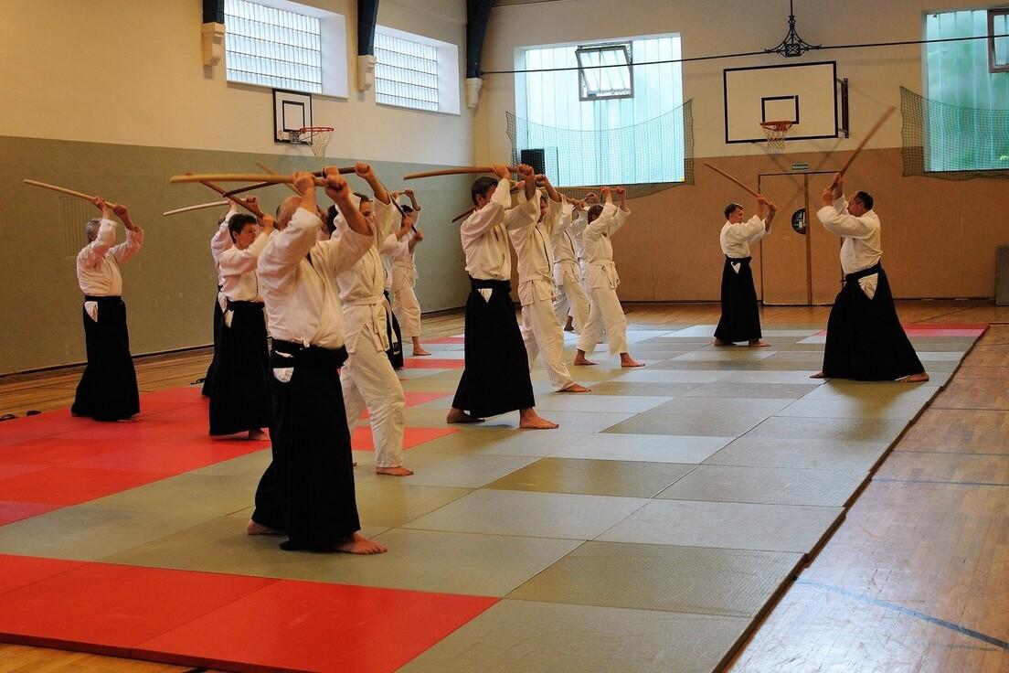 Gruppentraining in einer Kampfsporthalle: Mitglieder in traditioneller Kleidung &uuml;ben mit Holzschwertern auf Matten.