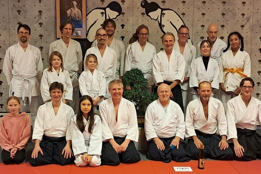 Gruppe von 20 Personen in wei&szlig;en Judogis, posierend in einem Dojo mit Pflanzen und einem Wandbild im Hintergrund.