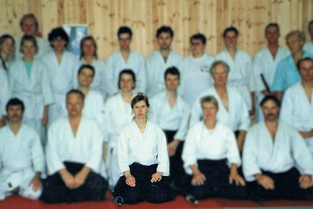 Gruppe von Aikido-Praktizierenden in wei&szlig;en Gew&auml;ndern, sitzend und stehend, vor einer Holztafel im Hintergrund.