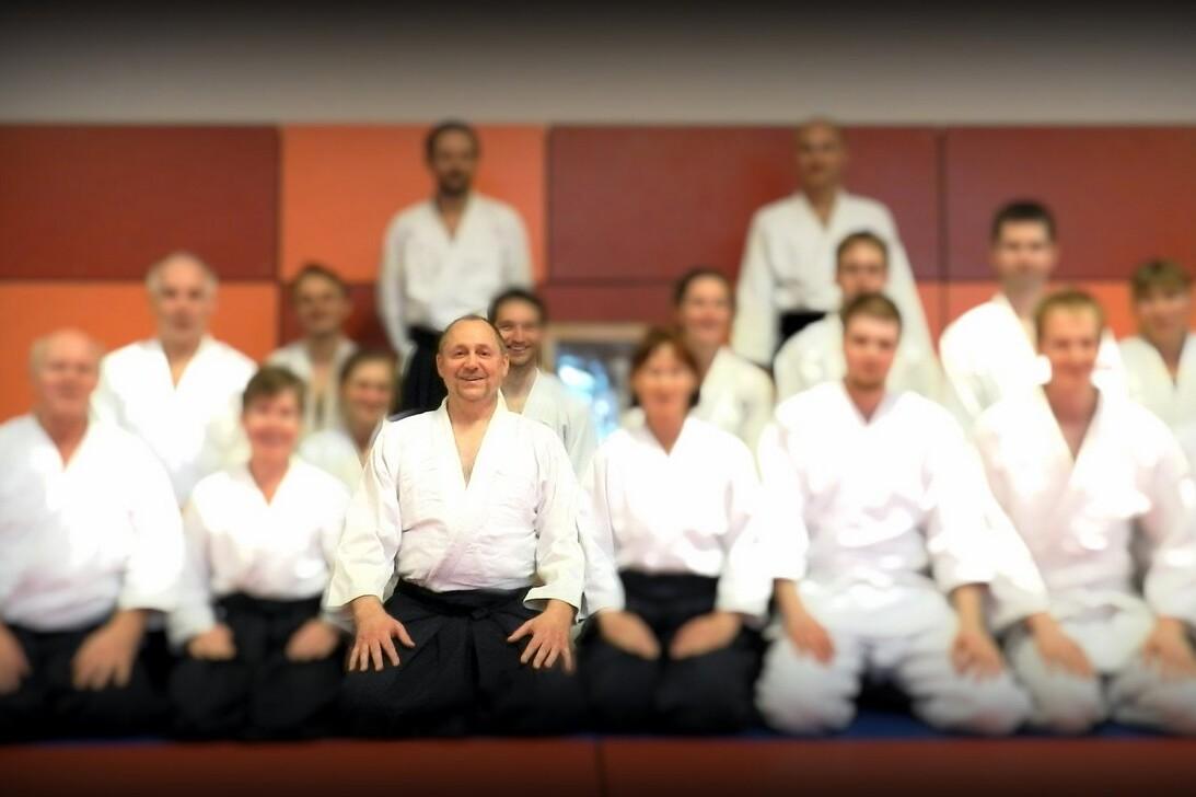 Gruppe von Aikido-Praktizierenden in wei&szlig;en Judokas und schwarzen Hosen, sitzend vor einer bunten Wand.