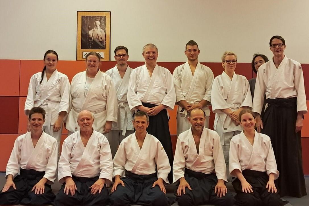 Gruppe von zw&ouml;lf Personen in wei&szlig;en Martial-Arts-Anz&uuml;gen, sitzend und stehend, vor einer querfarbenen Wand.