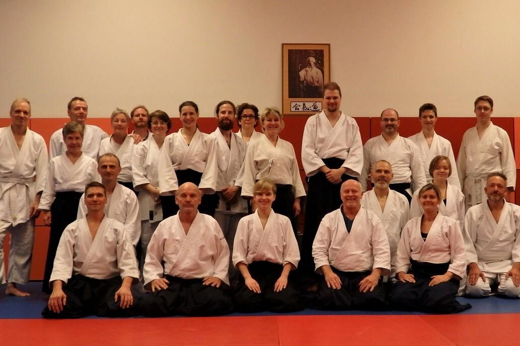 Gruppenfoto von Aikido-Praktizierenden in wei&szlig;en und schwarzen Anz&uuml;gen auf einer blauen und roten Matte.