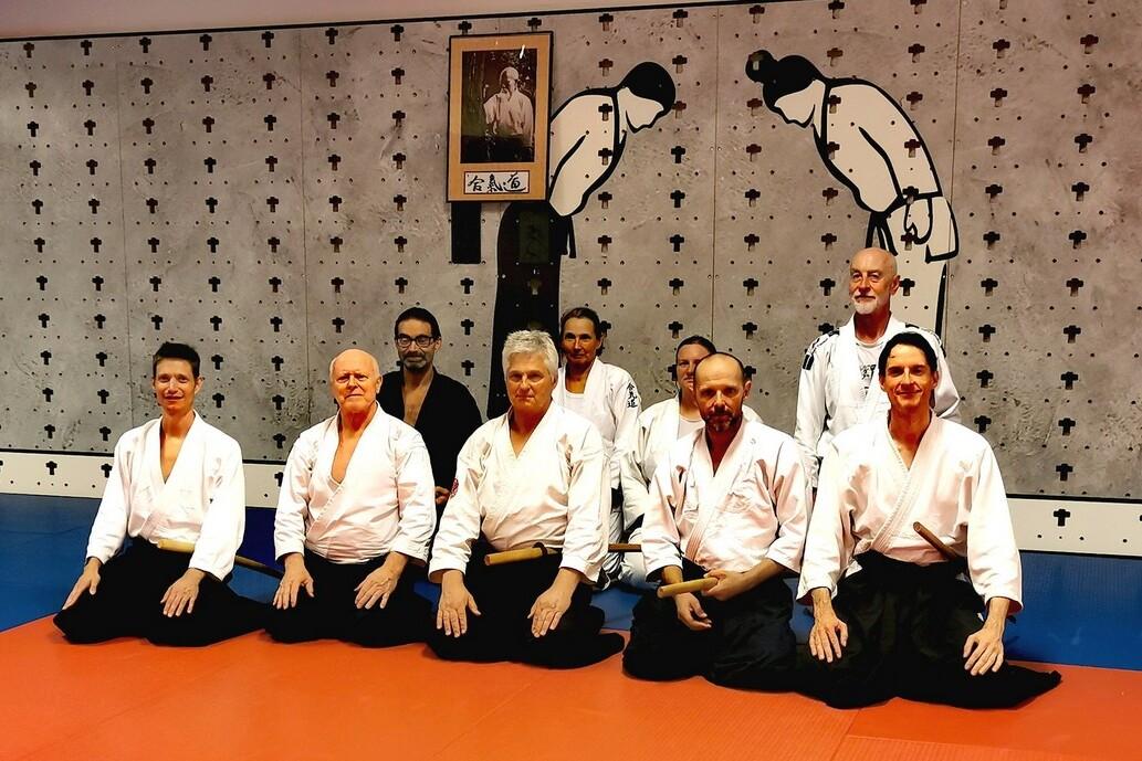 Gruppe von acht Personen im Sitz, in traditionellen Uke-Gi, in einem Aikido-Dojo mit Wandkunst im Hintergrund.