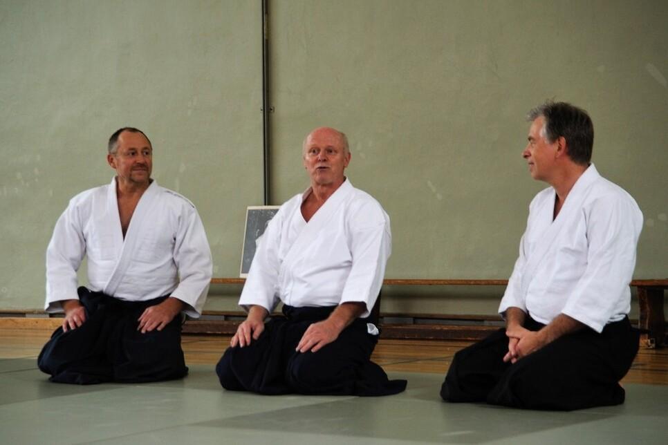 Drei M&auml;nner in traditioneller Aikido-Gewandung sitzen auf einer Matte in einem Trainingsraum und unterhalten sich.