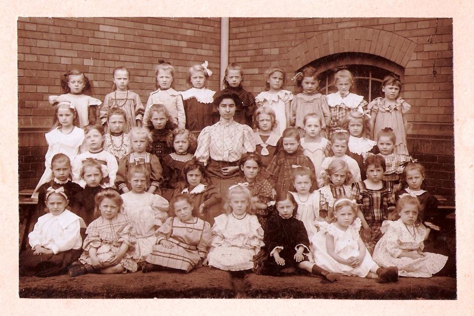 Gruppenbild von 34 M&auml;dchen in historischen Kleidern, umgeben von Ziegelmauer, einige tragen Haarschleifen.