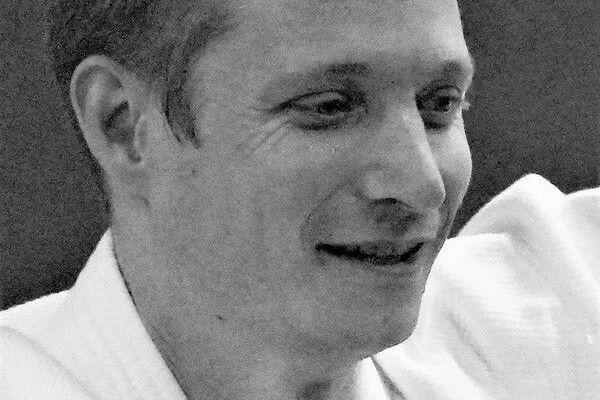 Lächelnder Mann in weißem Judogi auf einem dunklen Hintergrund, ein unbeschwertes Lächeln auf seinem Gesicht.
