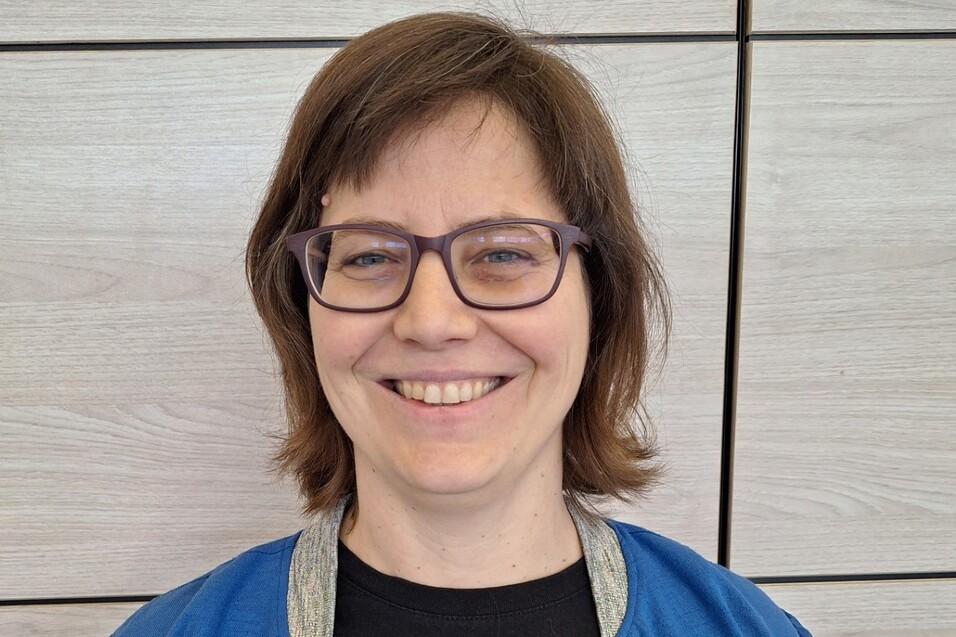 Frau mit Brille tr&auml;gt ein schwarzes T-Shirt mit "V. HAMBURG BASKETBALL" und eine blaue Jacke, l&auml;chelt vor Holzpaneelen.