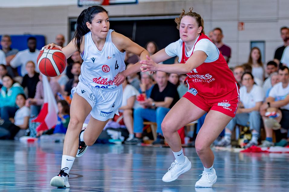 Zwei Basketballspielerinnen dribbeln den Ball auf dem Spielfeld, während Zuschauer im Hintergrund zuschauen.