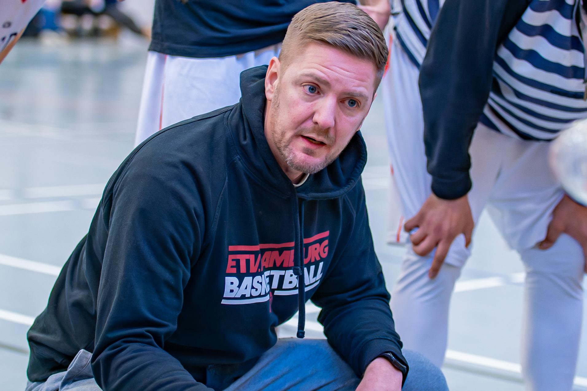 Basketballtrainer kniet im Gespr&auml;ch mit Spielern, w&auml;hrend sie sich um ihn versammeln und Waterflaschen halten.