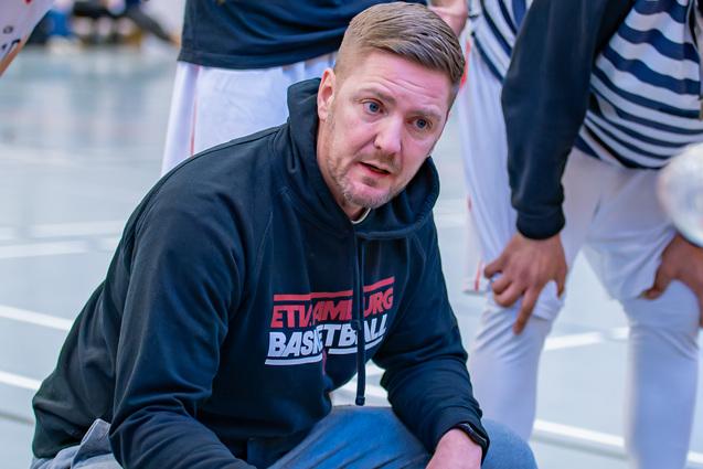 Basketballtrainer kniet im Gespr&auml;ch mit Spielern, w&auml;hrend sie sich um ihn versammeln und Waterflaschen halten.