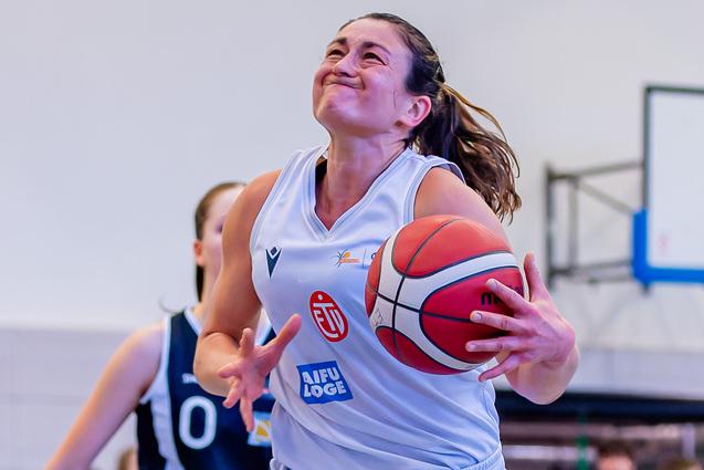 Basketballspielerin mit Ball in der Hand springt zum Korb, w&auml;hrend eine Gegnerin sie verfolgt. Zuschauer im Hintergrund.