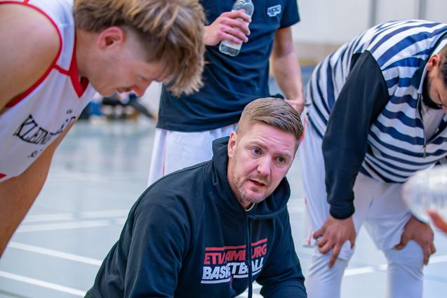 Basketballtrainer kniet im Gespr&auml;ch mit Spielern, w&auml;hrend sie sich um ihn versammeln und Waterflaschen halten.