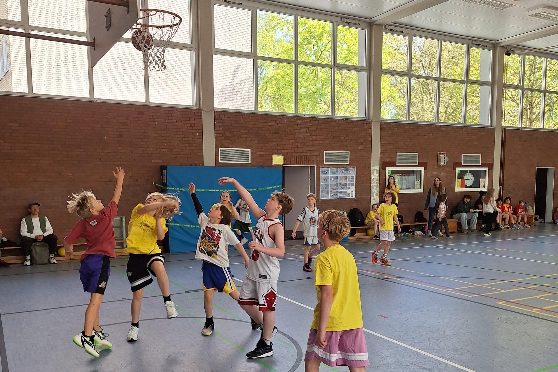 Mehrere Kinder spielen Basketball in einer Halle, w&auml;hrend Zuschauer auf B&auml;nken am Rand sitzen.