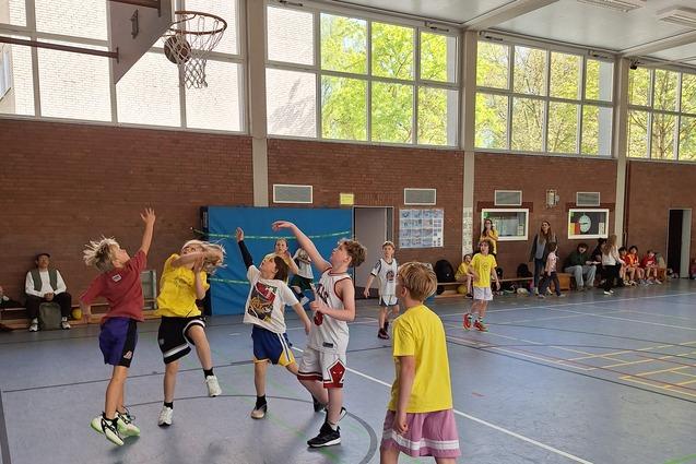 Mehrere Kinder spielen Basketball in einer Halle, w&auml;hrend Zuschauer auf B&auml;nken am Rand sitzen.
