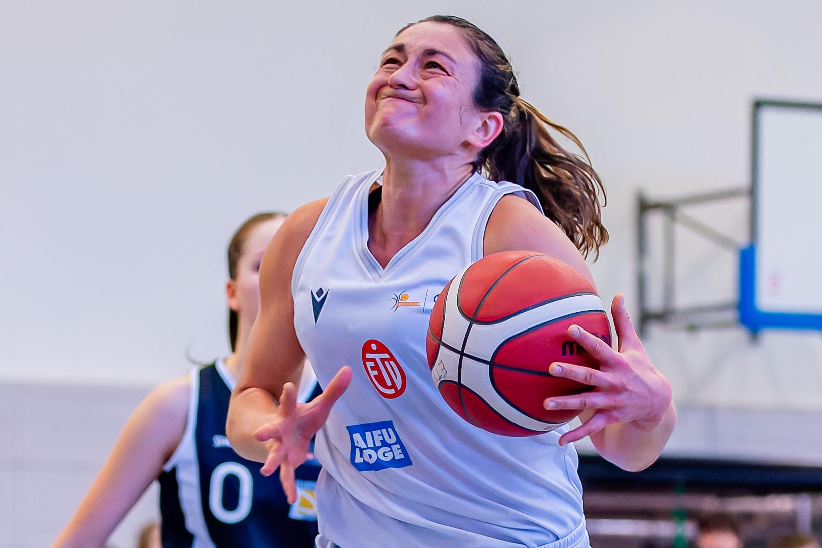 Basketballspielerin mit Ball in der Hand springt zum Korb, w&auml;hrend eine Gegnerin sie verfolgt. Zuschauer im Hintergrund.