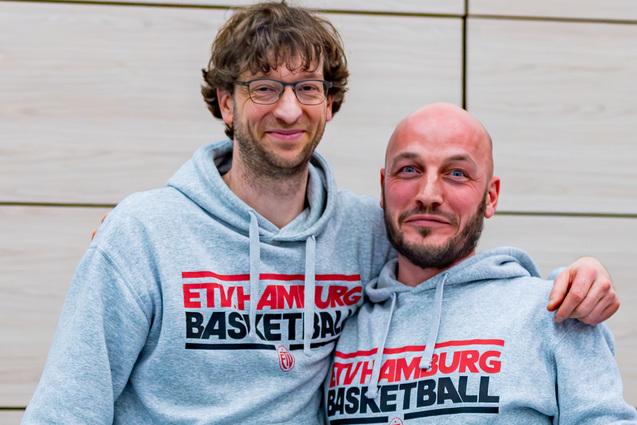 Zwei Männer in grauen Sweatshirts mit "EHTY AMBURG BASKETBALL" posieren lächelnd vor einer hellen Wand.