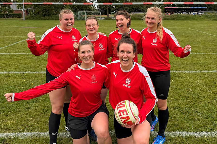 Sechs Frauen in roten Sporttrikots posieren fr&ouml;hlich auf einem Rasenfeld, einige halten einen Rugbyball.