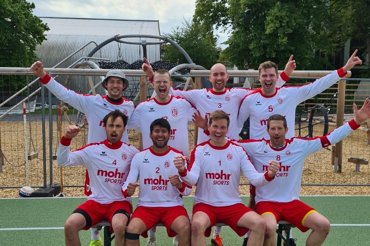 Gruppenzusammenstellung von acht Fu&szlig;ballspielern in roten und wei&szlig;en Trikots, l&auml;chelnd auf einem Sportplatz.