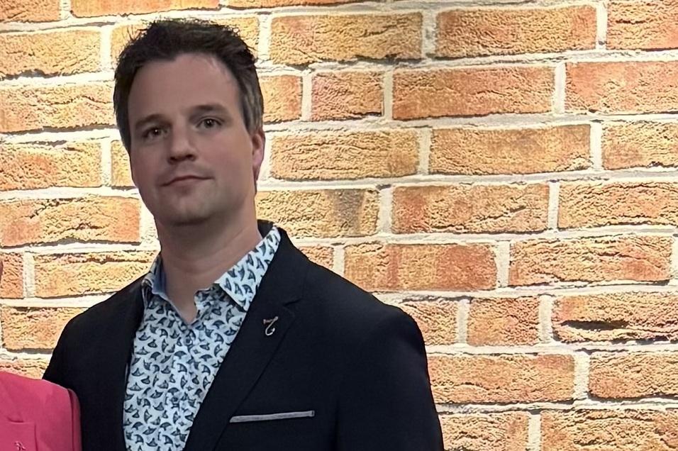 Foto Florian Mann in einem dunklen Blazer mit gemusterter Bluse vor einer Backsteinwand, hält ein Dokument in der Hand.