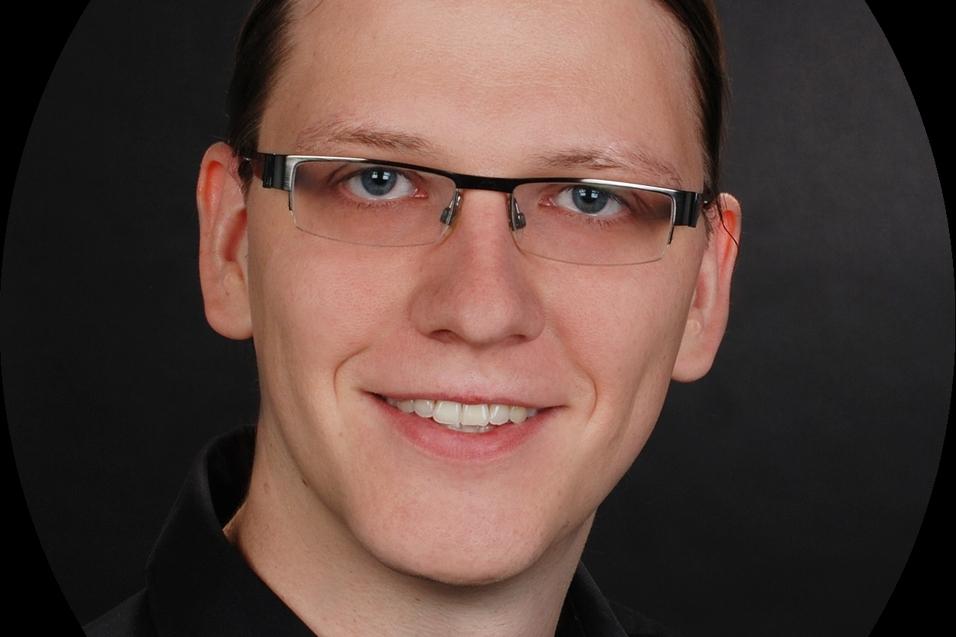 Porträt eines Mannes mit Brille und langen, hellbraunen Haaren vor einem dunklen Hintergrund. Er lächelt freundlich.