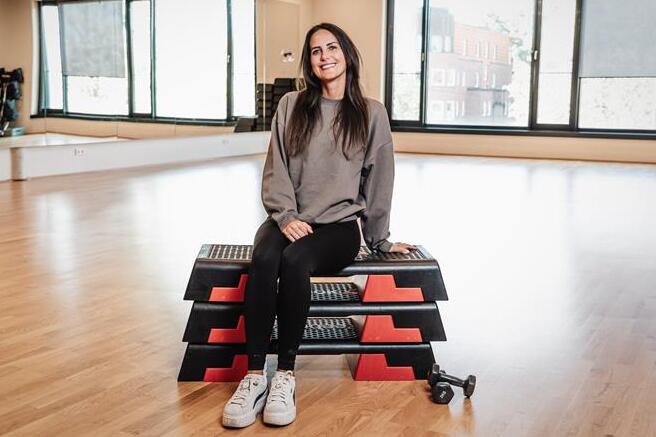 Frau sitzt auf einem schwarzen Step-Brett in einem Fitnessstudio, lächelt und trägt sportliche Kleidung.