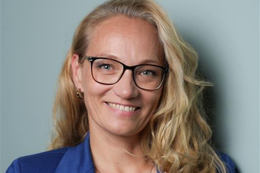 Porträt einer lächelnden Frau mit blonden, lockigen Haaren, Brille und blauem Blazer vor einer grauen Wand.