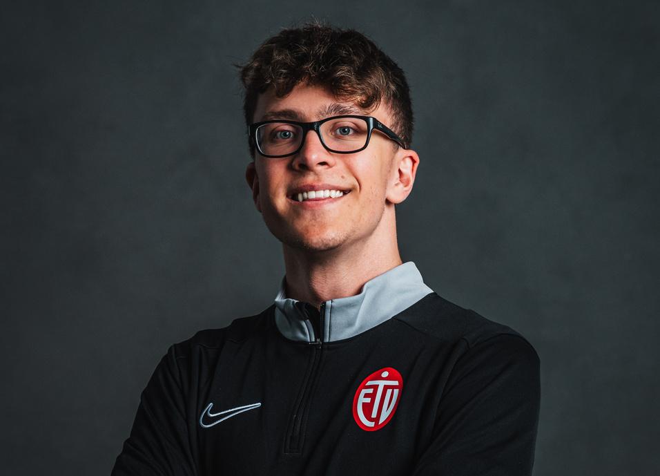 Junger Mann mit Brille in schwarzem Sportpullover, l&auml;chelt und hat die Arme vor der Brust verschr&auml;nkt, vor neutralem Hintergrund.