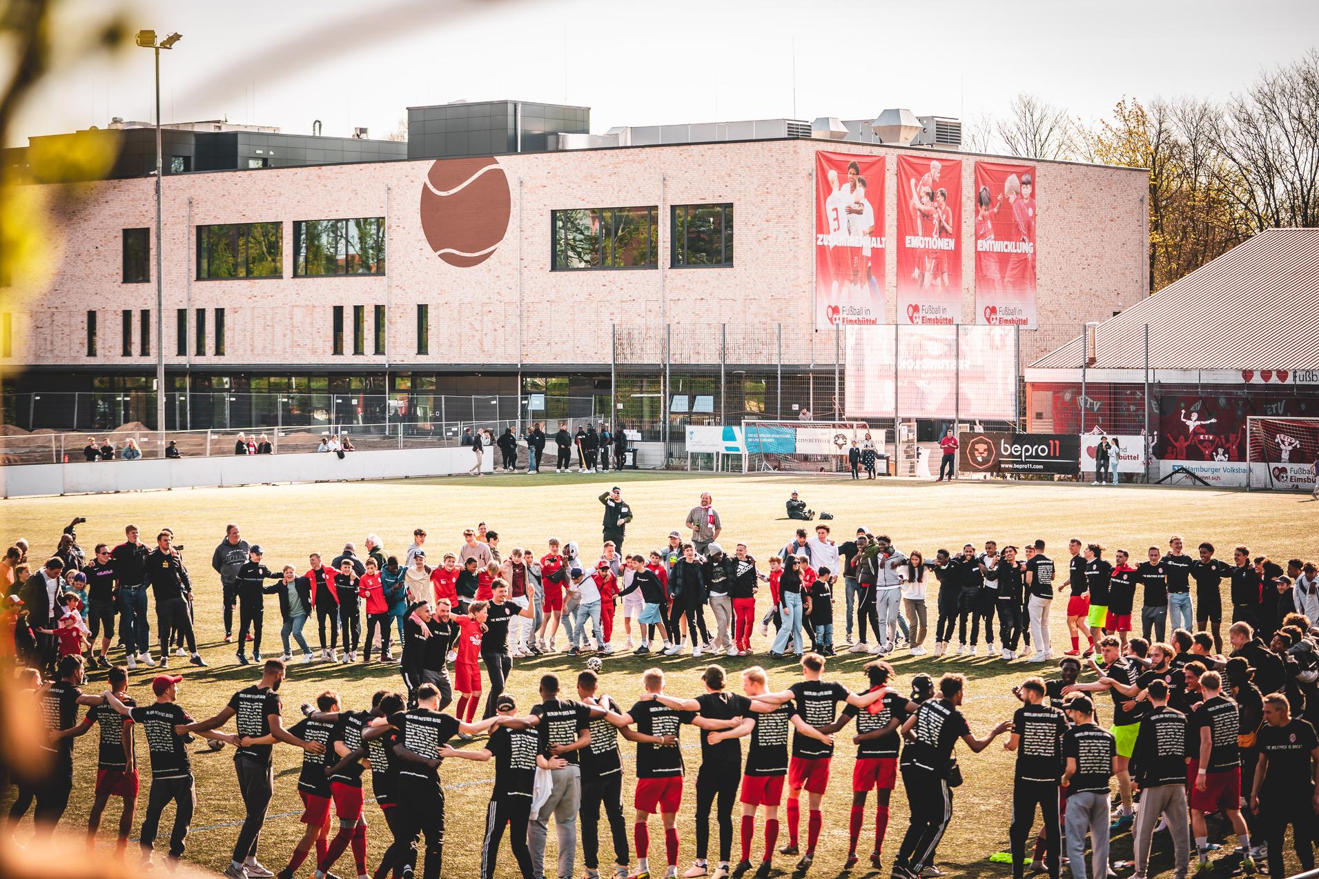 Gruppierung von Spielern und Zuschauern auf einem Sportplatz, um einen Kreis mit dem Vereinsgeb&auml;ude im Hintergrund zu bilden.