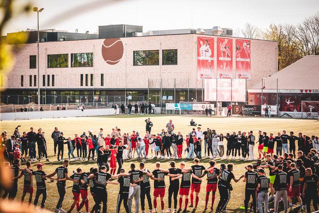 Gruppierung von Spielern und Zuschauern auf einem Sportplatz, um einen Kreis mit dem Vereinsgeb&auml;ude im Hintergrund zu bilden.