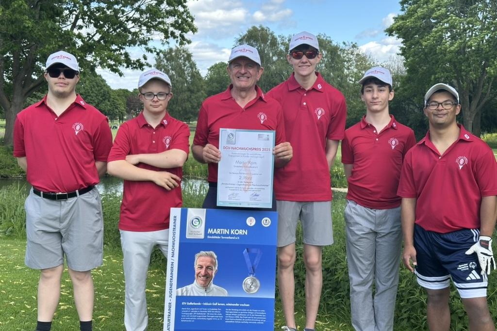 Sechsk&ouml;pfige Gruppe in roten Poloshirts posiert neben einer Urkunde und einem Informationsschild auf einem Golfplatz.