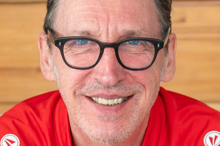 Porträt eines lächelnden Mannes in rot-blauem Outfit mit Brille, vor einer Holzoberfläche.