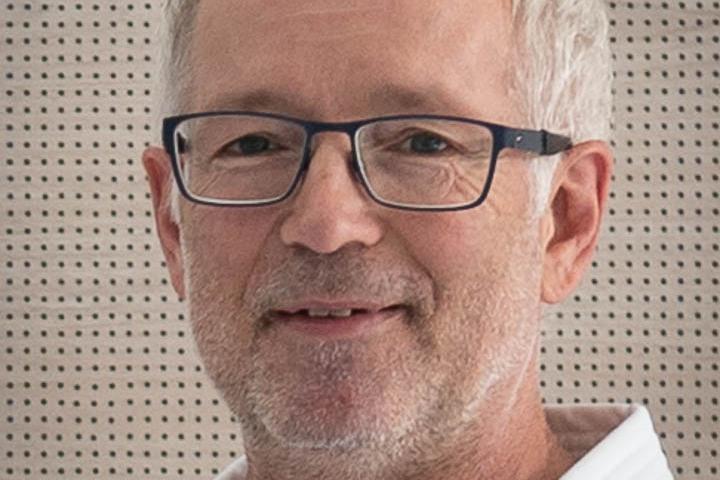 Lächelnder Mann mit Brille und grauen Haaren in einem weißen Karate-Gi vor einer hellen, strukturierten Wand.