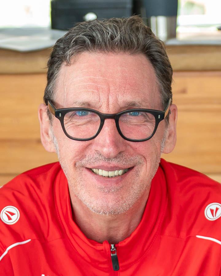 Porträt eines lächelnden Mannes in rot-blauem Outfit mit Brille, vor einer Holzoberfläche.