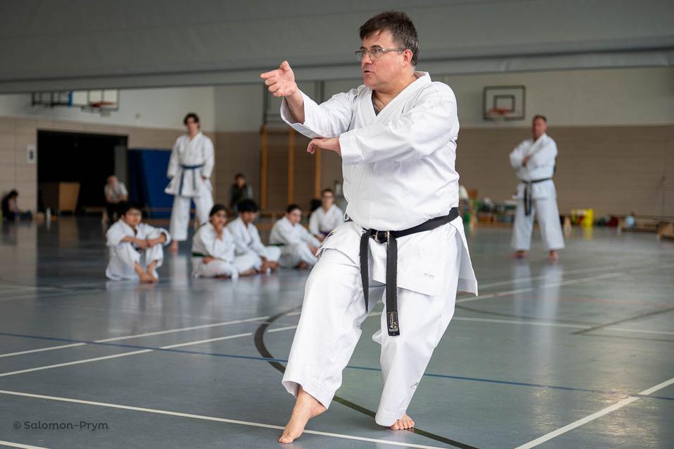 Ein Karateka in Wei&szlig; f&uuml;hrt einen Angriff im Dojo aus, w&auml;hrend andere Trainingsteilnehmer zuschauen.
