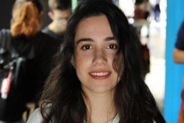 Porträt einer jungen Frau mit langen, dunklen Haaren, die in einer belebten Umgebung lächelt. Unschärfe im Hintergrund.