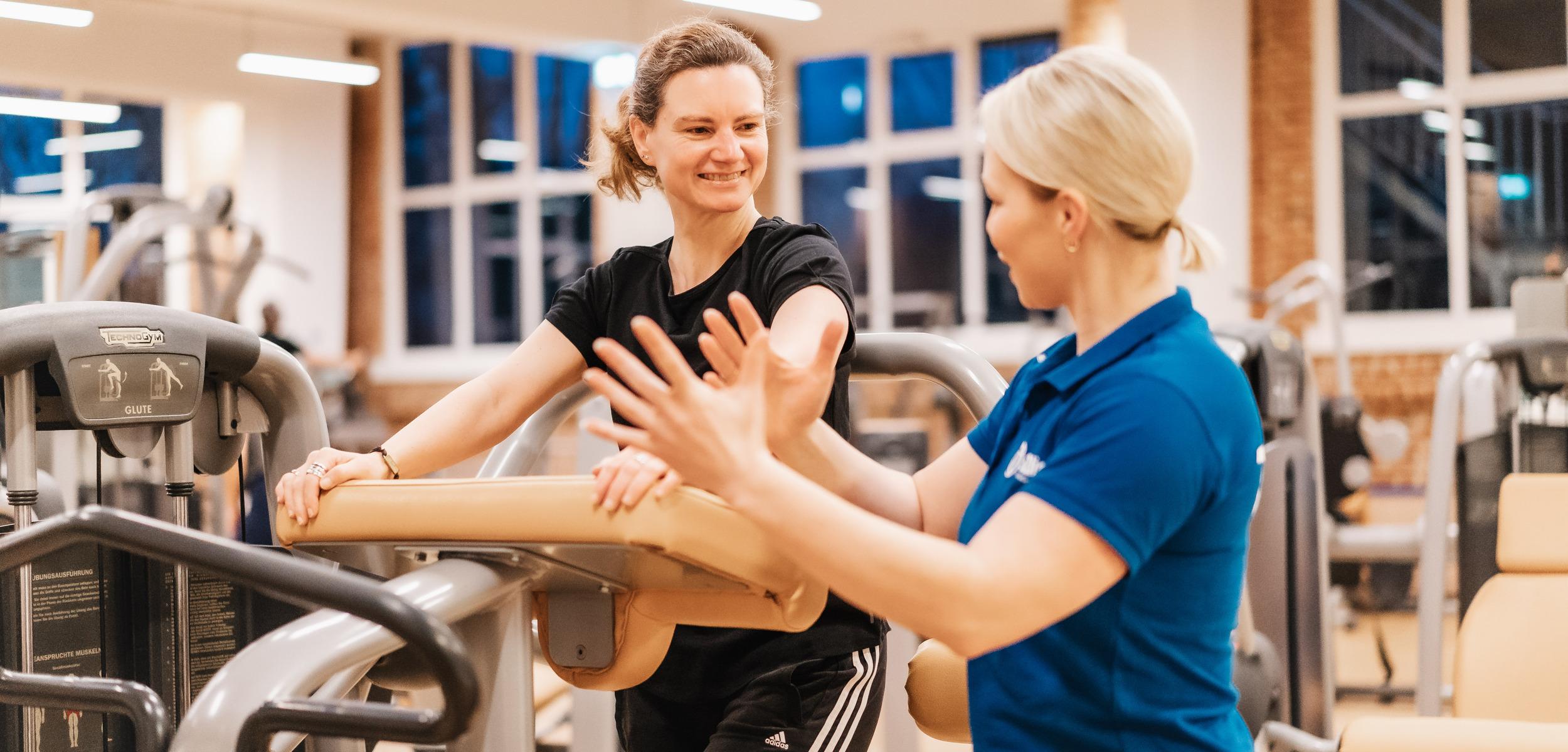 Zwei Frauen in Sportkleidung unterhalten sich in einem Fitnessstudio, w&auml;hrend eine an einem Trainingsger&auml;t steht.