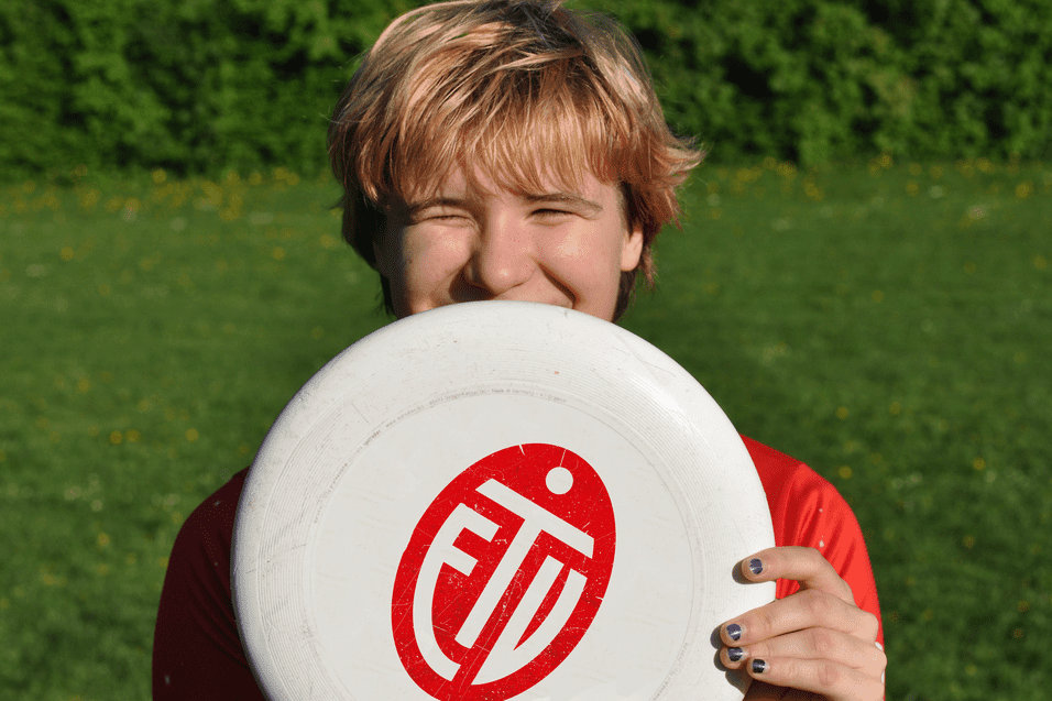 L&auml;chelnder junger Mann h&auml;lt eine Frisbee vor seinem Gesicht, auf einer gr&uuml;nen Wiese mit Bl&uuml;ten.