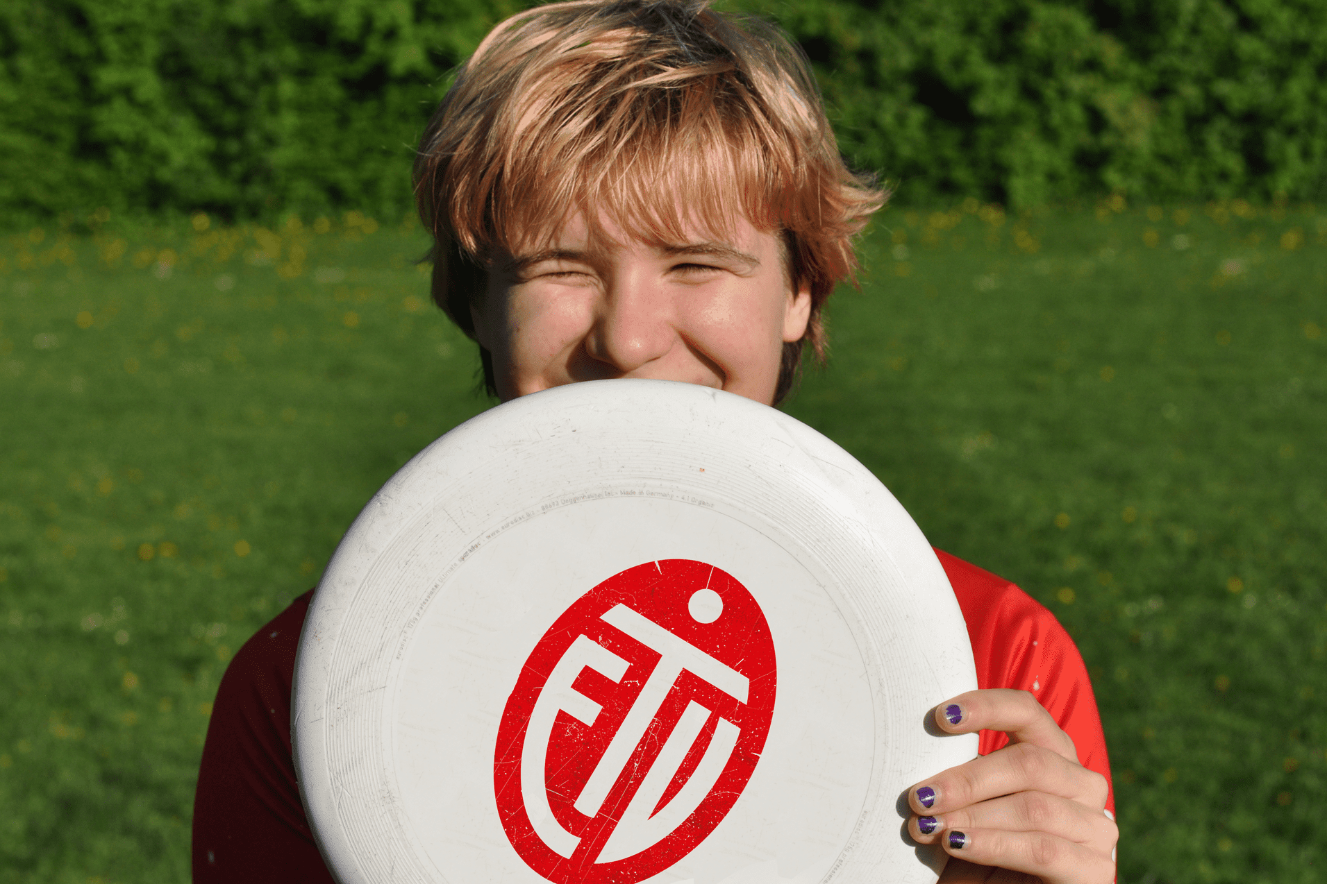 Lächelnder junger Mann hält eine Frisbee vor seinem Gesicht, auf einer grünen Wiese mit Blüten.