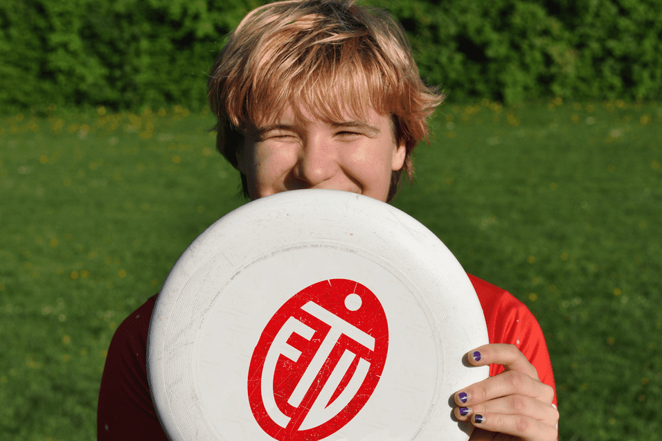 L&auml;chelnder junger Mann h&auml;lt eine Frisbee vor seinem Gesicht, auf einer gr&uuml;nen Wiese mit Bl&uuml;ten.