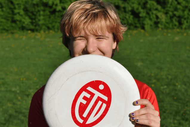 Lächelnder junger Mann hält eine Frisbee vor seinem Gesicht, auf einer grünen Wiese mit Blüten.