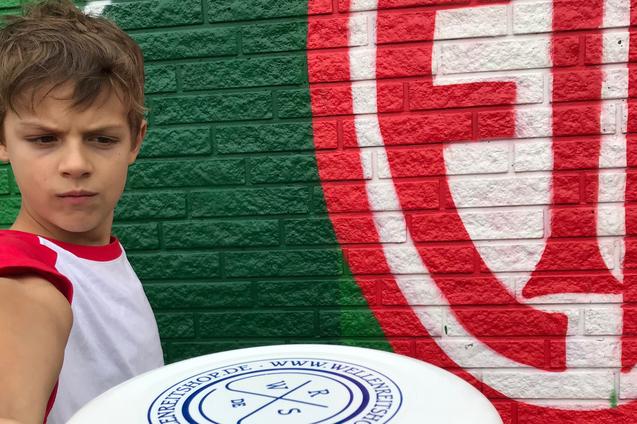 Ein Junge h&auml;lt eine Frisbee in Richtung Kamera, vor einer bunten Wand mit gr&uuml;nen und roten Mustern.