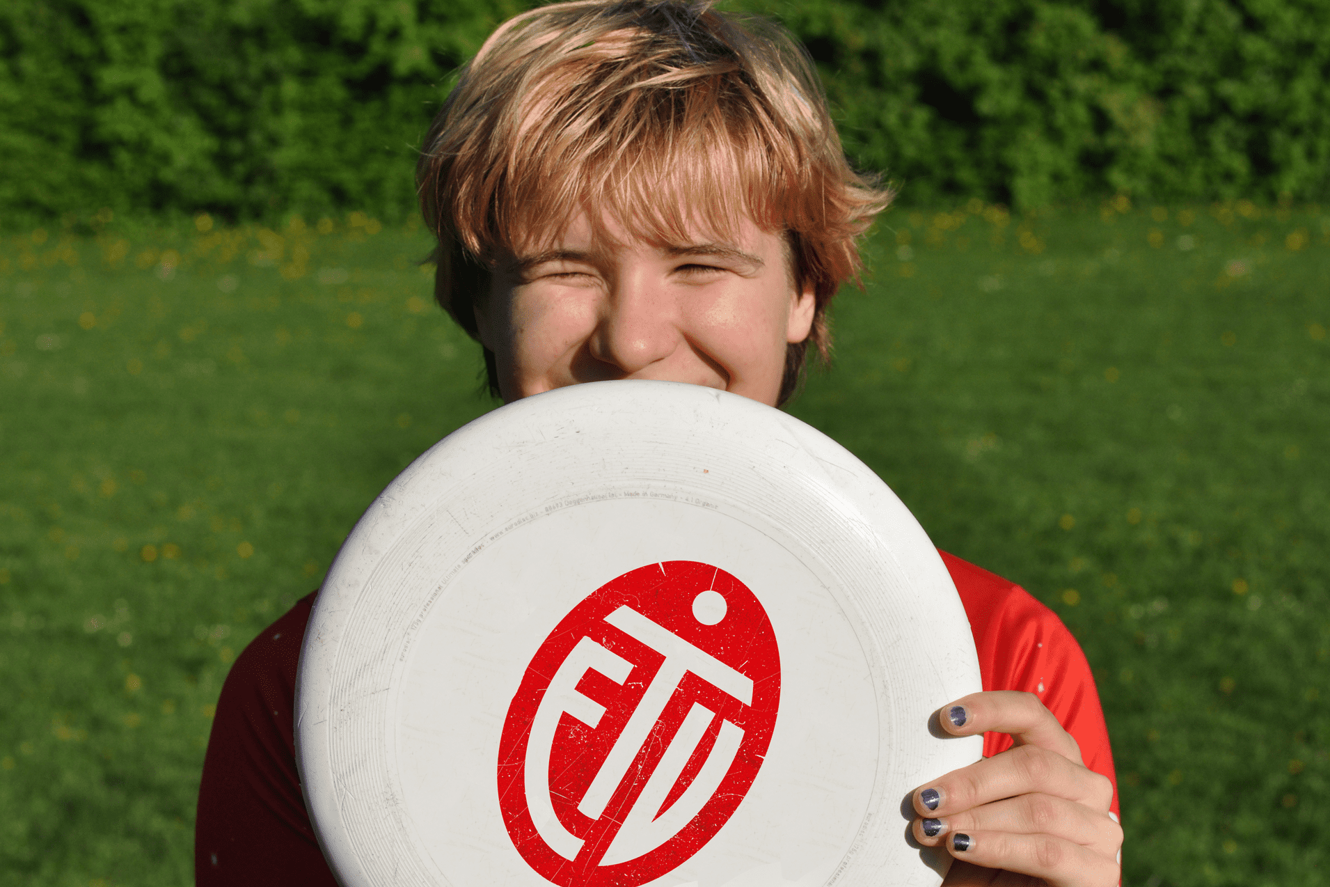 Lächelnder junger Mann hält eine Frisbee vor seinem Gesicht, auf einer grünen Wiese mit Blüten.