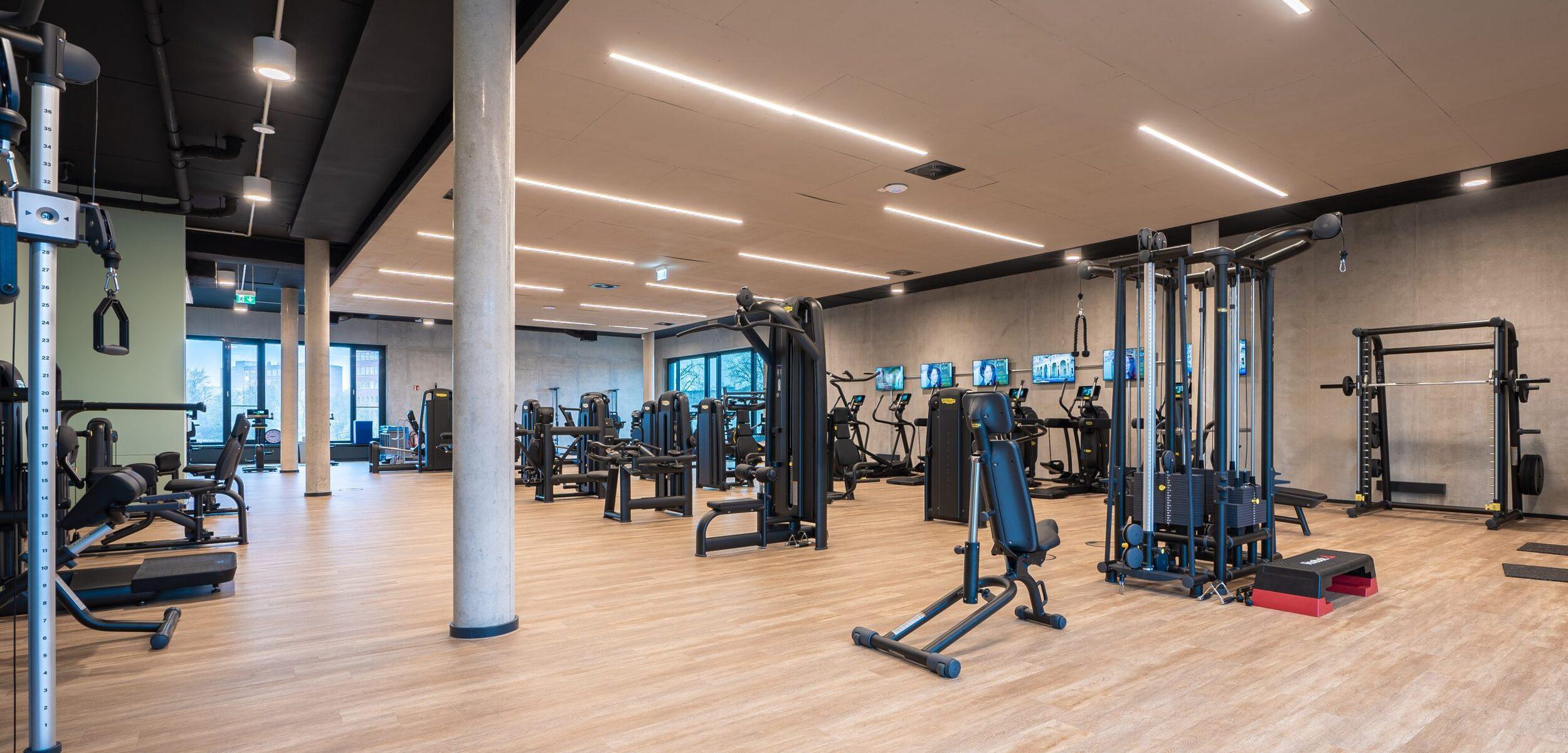 Heller Fitnessraum mit verschiedenen Trainingsger&auml;ten, gro&szlig;en Fenstern und einem Holzfu&szlig;boden.