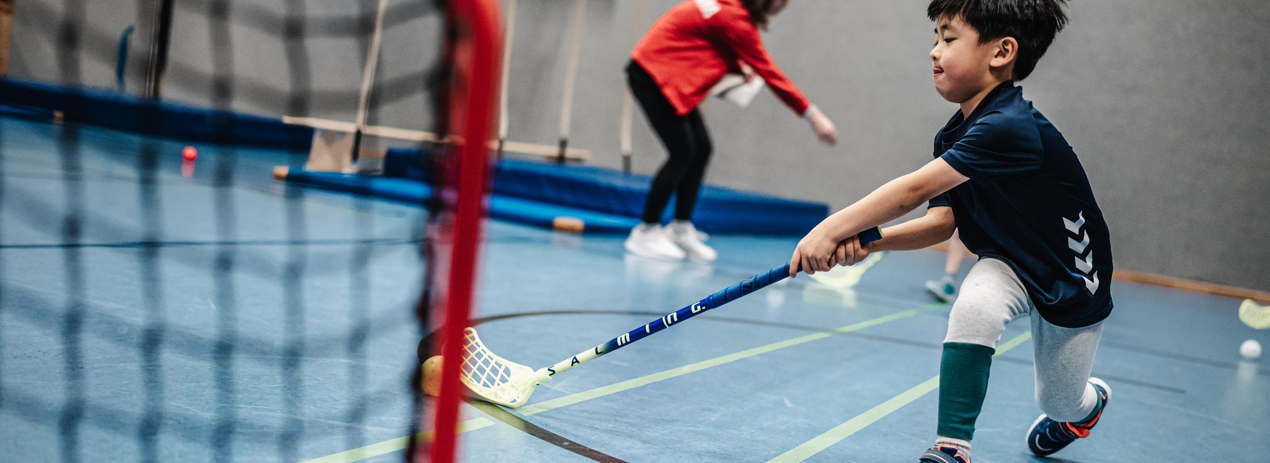 Junge mit einem Stock im Hallen-Sportraum, w&auml;hrend er auf ein Tor zielt. Im Hintergrund eine Trainerin.
