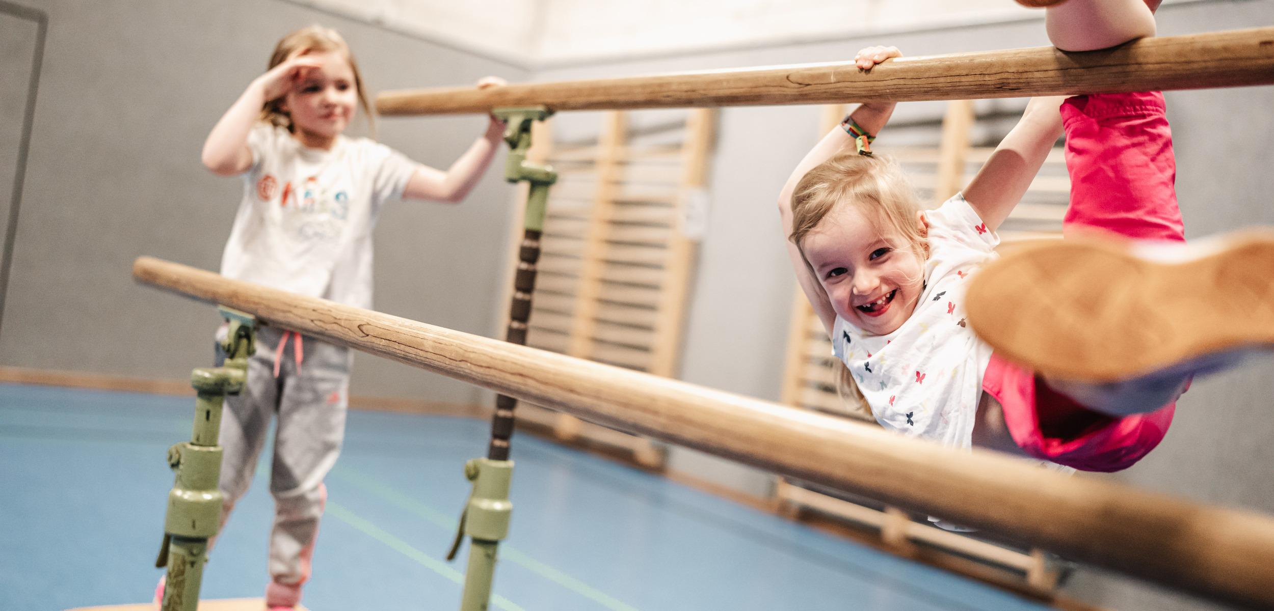 Zwei Kinder spielen in einer Sporthalle an einem Barren, eines h&auml;ngt l&auml;chelnd, das andere steht daneben.